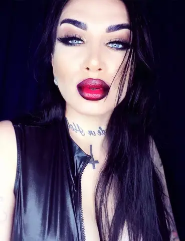 Woman with dark hair and red lipstick wearing a black sleeveless top against a dark background.