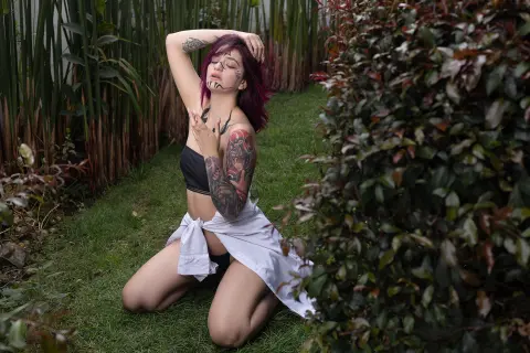 Tattooed woman in a black crop top and white wrap sitting on grass in a lush garden setting.