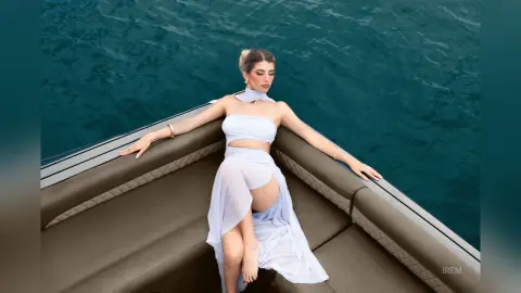 Elegant woman in a white flowing summer outfit relaxing on a luxury boat in turquoise ocean water.