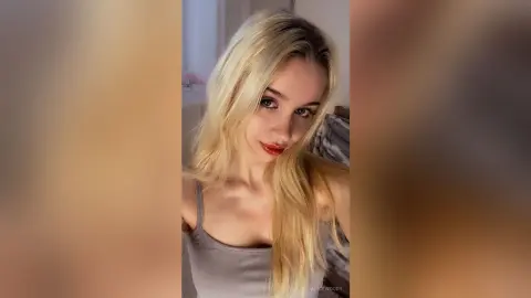 Blonde woman in a grey tank top posing with a soft, natural look in a bright indoor setting.