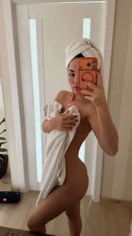 Young woman in a white spa towel taking a mirror selfie in a bright bathroom setting.