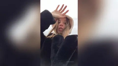 Blonde woman in a cozy black faux fur coat and white hoodie posing outdoors against a soft, bright sky.