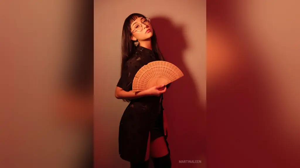 MartinaLeen β Stylish woman in a black dress holding a wooden hand fan against a warm, moody red studio background.
