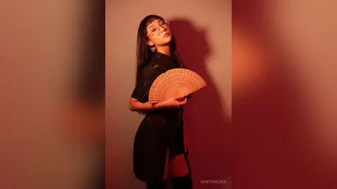 Stylish woman in a black dress holding a wooden hand fan against a warm, moody red studio background.