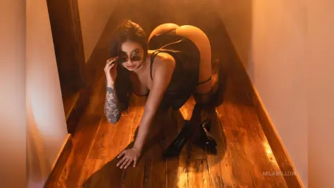 MilaWillow crawls on a wooden floor in a dark hallway, wearing black sunglasses and a black top that shows her bare ass.