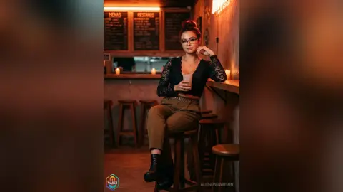Stylish woman in a black lace top and khaki trousers sitting at a moody, dimly lit cafe bar.