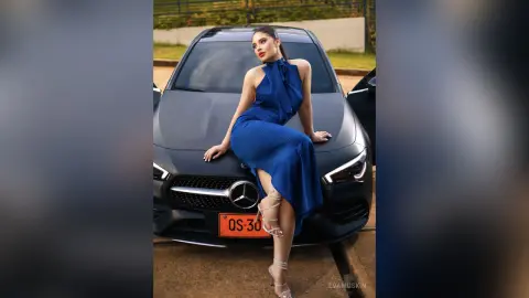 Elegant woman in a royal blue halter dress leaning on a black Mercedes parked outdoors in soft daylight.