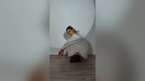 Stylish woman in an oversized beige shirt and black boots posing in a minimalist white room.