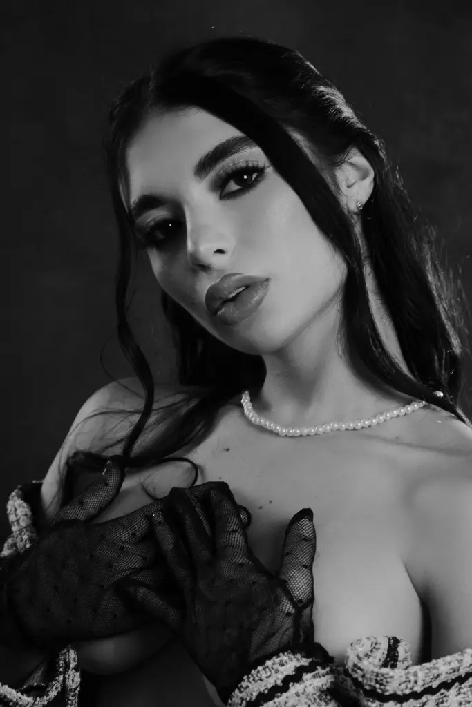 Glamorous woman with long dark hair in a black off-the-shoulder top posing against a grey studio background.