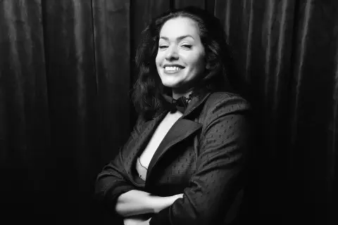 Smiling woman in a dark blazer posing against a black velvet curtain in a moody black and white portrait.