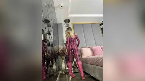 NicoleBella stands in a grey studio wearing a pink patterned bodysuit and matching pink boots, showing her back and blonde hair.