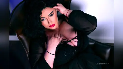 AdiraMaeven poses in a black top with a deep neckline, running her hand through her dark wavy hair while sitting in a dark leather chair.