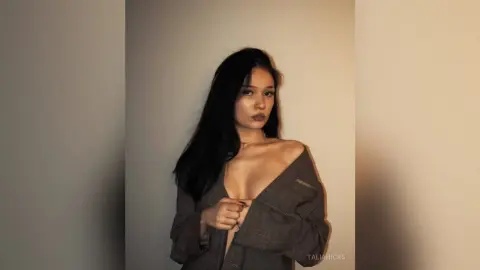 Young woman in a grey plaid blazer posing against a neutral wall in moody, warm lighting.