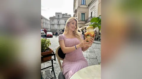 Smiling blonde woman in a pink ribbed dress enjoying a refreshing drink at an outdoor European cafe.