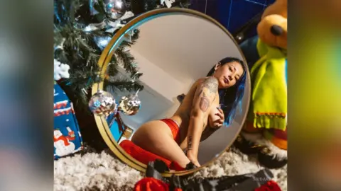 Blue-haired woman in red lingerie posing in a mirror near a festive Christmas tree and teddy bear.