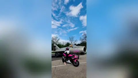 Woman in black riding gear sitting on a pink sport motorcycle in front of a vintage fighter jet under a blue sky.