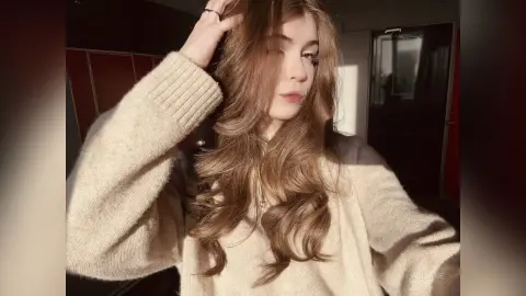 Young woman with wavy brown hair wearing a cozy beige sweater in warm sunlight.