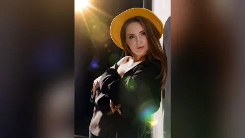 Stylish woman in a black top and yellow wide-brimmed hat posing in dramatic sunlight.