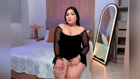 Young woman in a black sheer bodysuit sitting on a bed in a bright, modern bedroom.