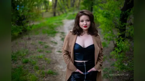 Smiling woman in a black leather corset and tan coat walking on a forest path during golden hour.