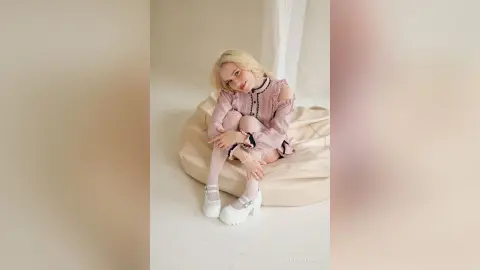LynGoodhart sits on a beige beanbag wearing a pink ruffled dress and white platform shoes.