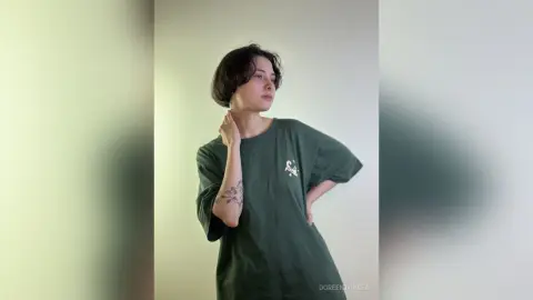 Young woman with short dark hair wearing a dark green graphic t-shirt in a minimalist indoor setting.