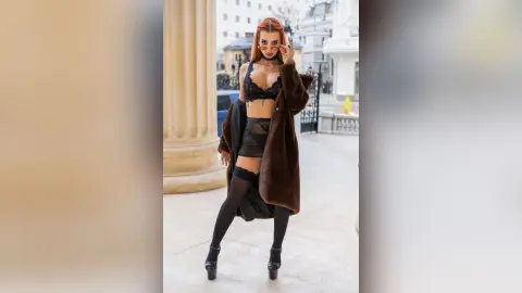 DeviNessa stands outdoors in a brown fur coat, black lace bra, and black micro skirt with black thigh-high stockings.