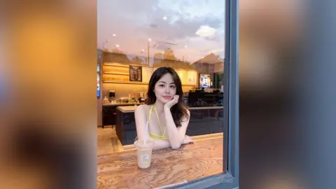 Young woman in a yellow tank top relaxing at a cafe window with an iced coffee during sunset.