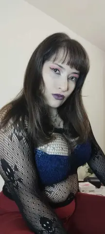 Woman with dark bangs wearing a blue lace bra and black fishnet top in a soft indoor setting.
