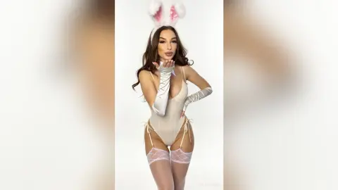 Beautiful woman in white bunny ears and satin bodysuit blowing a kiss against a white background.