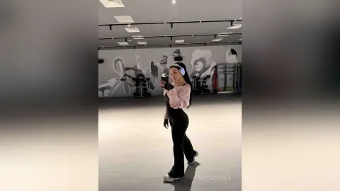 NicoleBella stands in a gym wearing a pink long-sleeve crop top, black leggings, and white headphones while taking a mirror selfie.