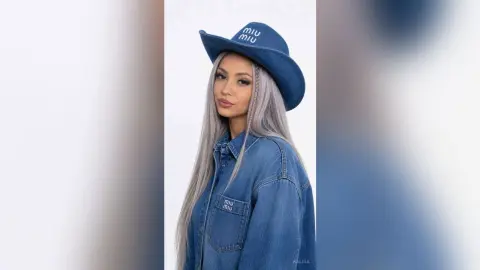 Kalira wears a blue denim shirt and a matching blue cowboy hat with white text, posing against a plain white background with her long grey hair down.