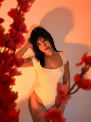 DapneOwen poses in a white bodysuit with her dark hair down, framed by red flowers against a warm background.