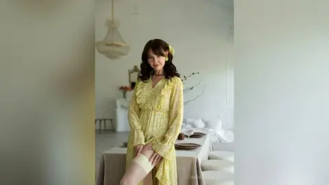 SophiaPanter poses in a yellow floral dress with long sleeves, standing in a bright room near a dining table.