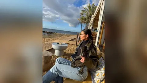 Stylish woman in a black turtleneck and olive jacket sitting at a beachside cafe under a cloudy sky.