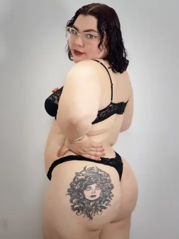 Curvy woman with glasses and Medusa tattoo wearing black lace lingerie against a plain white background.