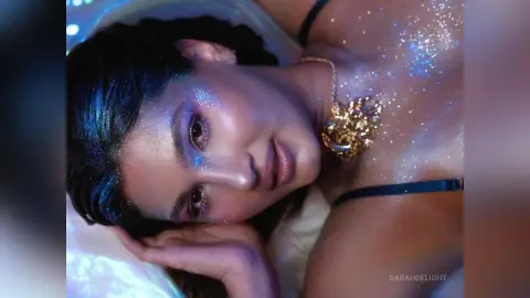 Glamorous woman with glittery skin and gold jewelry lying down in dreamy, ethereal lighting.