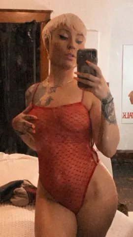 Blonde woman in a red textured bodysuit posing for a mirror selfie in a cozy bedroom setting.