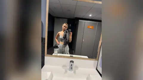 KeilaTouchton takes a mirror selfie in a grey crop top and sweatpants, holding her phone in a restroom setting.