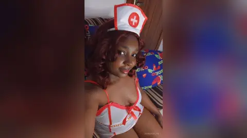 Smiling woman in a white and red nurse costume sitting on a bed in a cozy indoor setting.