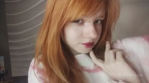 MozelleRozycki poses with her long red hair and red lipstick, wearing a white and pink knit sweater while looking at the camera.