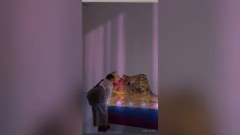 Woman in white blouse examining a large golden textured art installation in a minimalist gallery setting.