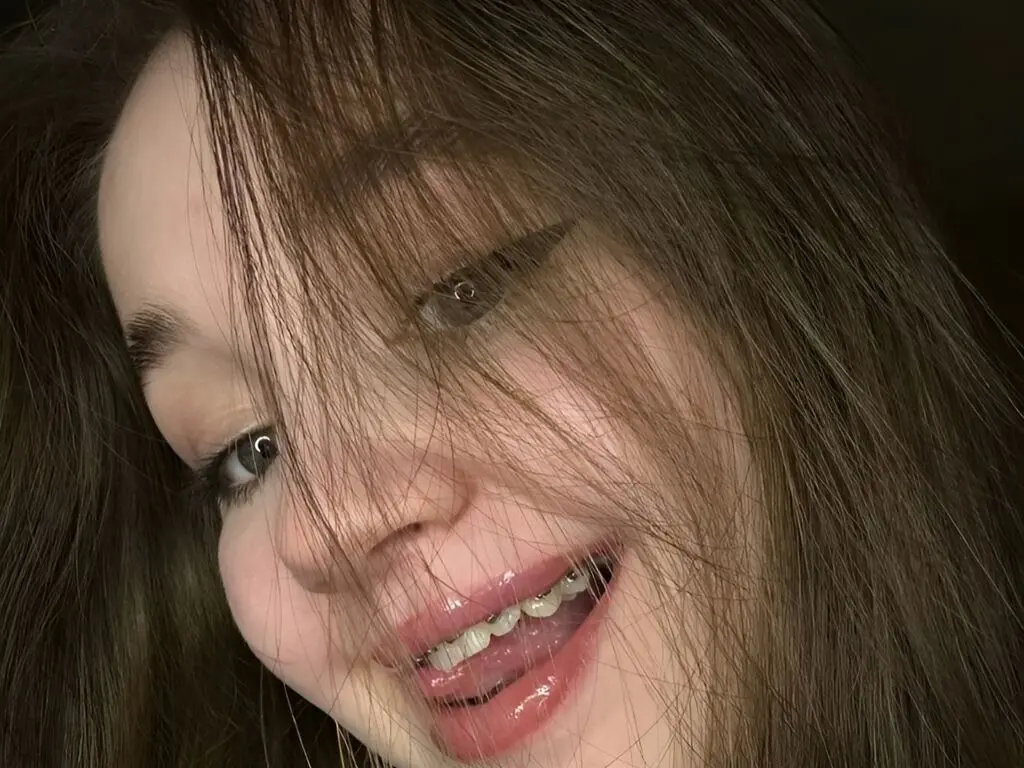 Close-up portrait of a smiling young woman with long brown hair and natural makeup in soft indoor lighting.
