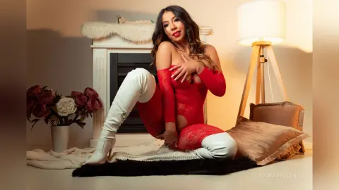 Glamorous woman in a red lace bodysuit and white thigh-high boots sitting on a cozy rug by a fireplace.