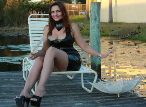 DominaPaulina sits on a white lounge chair by a lake wearing a tight black latex dress and holding a clear lace parasol.