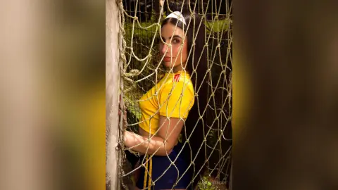 AlmaWhitmore poses behind a rope net wearing a yellow t-shirt and dark blue shorts with a white headband in her long dark hair.