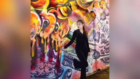 Young woman in a black jumpsuit standing before a vibrant, colorful street art mural in an urban setting.