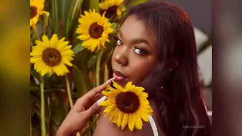 Beautiful young woman with long dark hair posing with bright sunflowers in a warm, sunny outdoor setting.