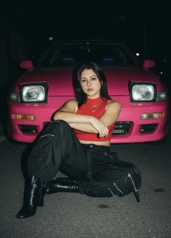 Stylish woman in a red crop top and black cargo pants sitting in front of a pink car at night.