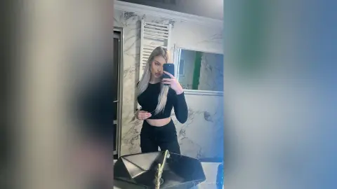 Young woman in a black long-sleeve crop top posing for a mirror selfie in a modern marble bathroom.